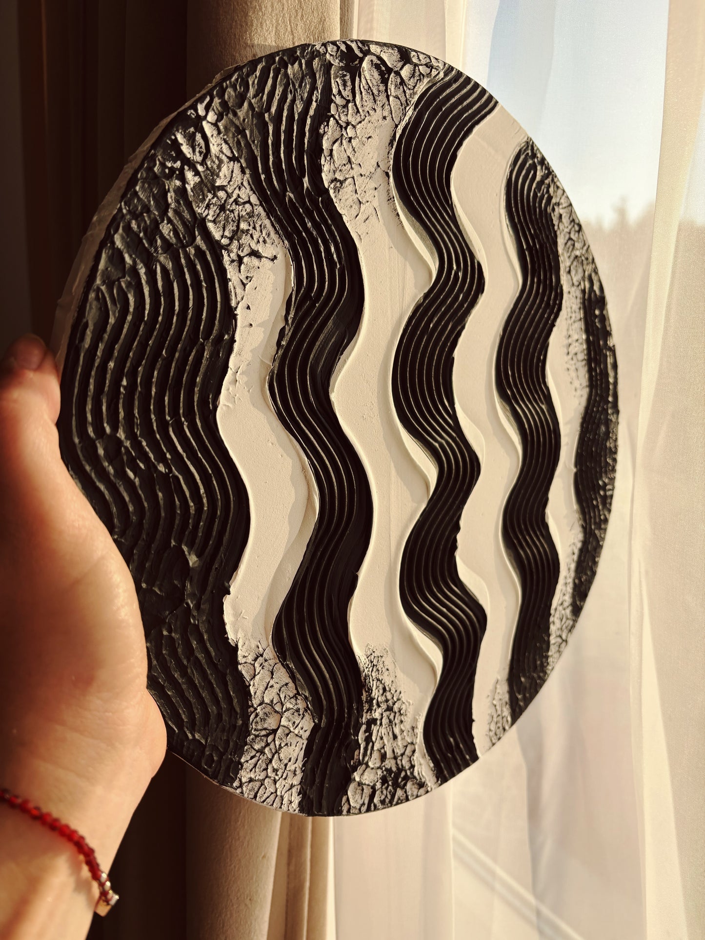 Round decorative wall art with wavy pattern held by a hand against a light curtain background