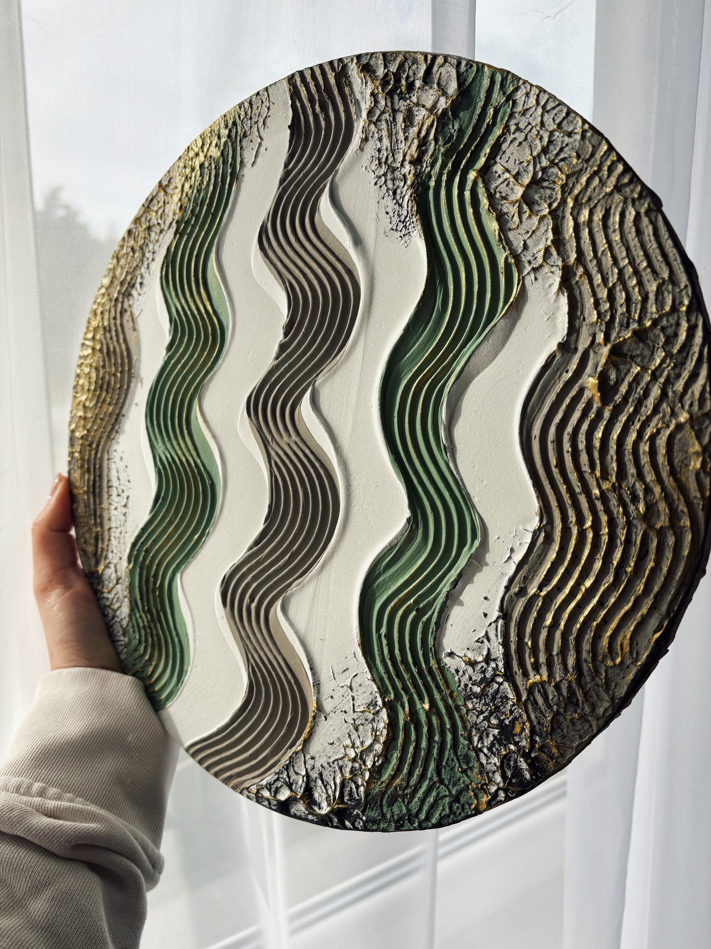 Round modern textured art with wavy green and brown design held by a person against a white curtain background