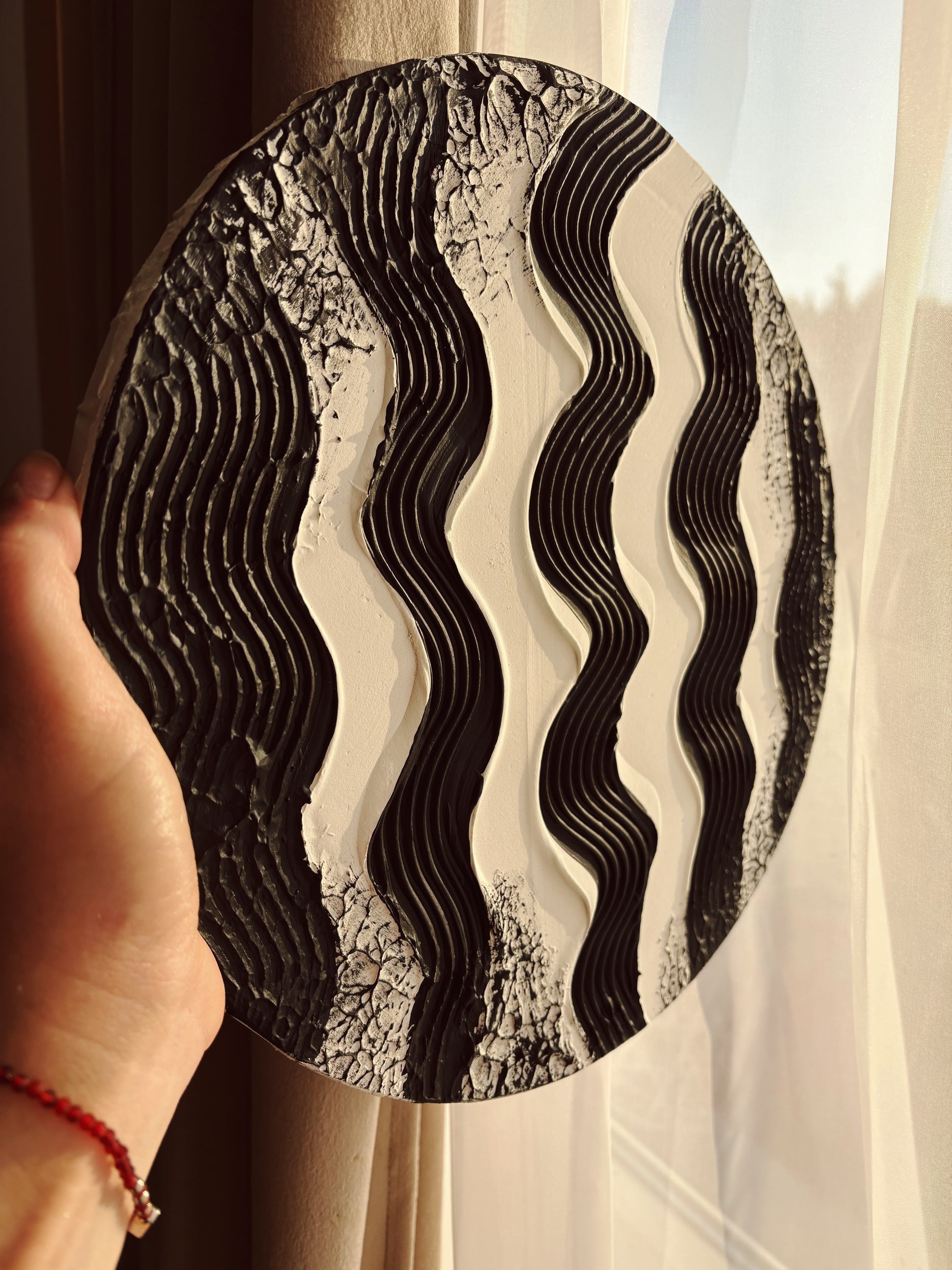 Round decorative wall art with wavy pattern held by a hand against a light curtain background