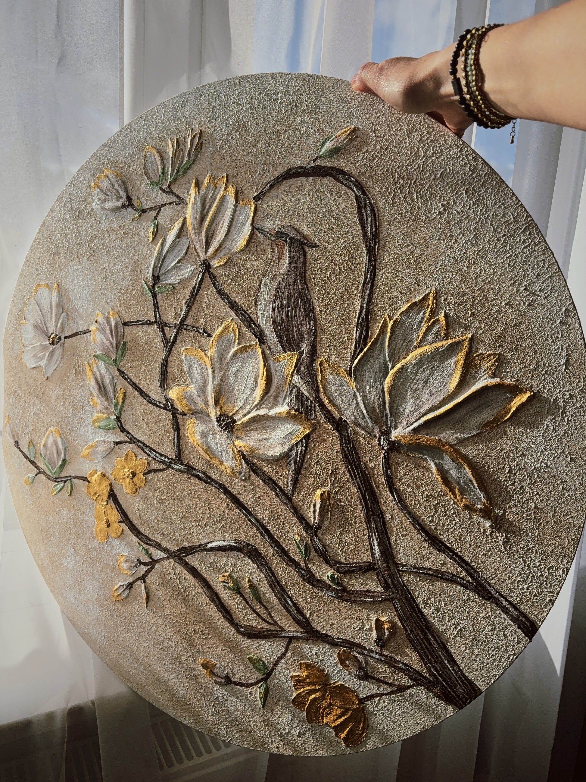 Round textured wall art with floral and bird design held by a hand against a light curtain background