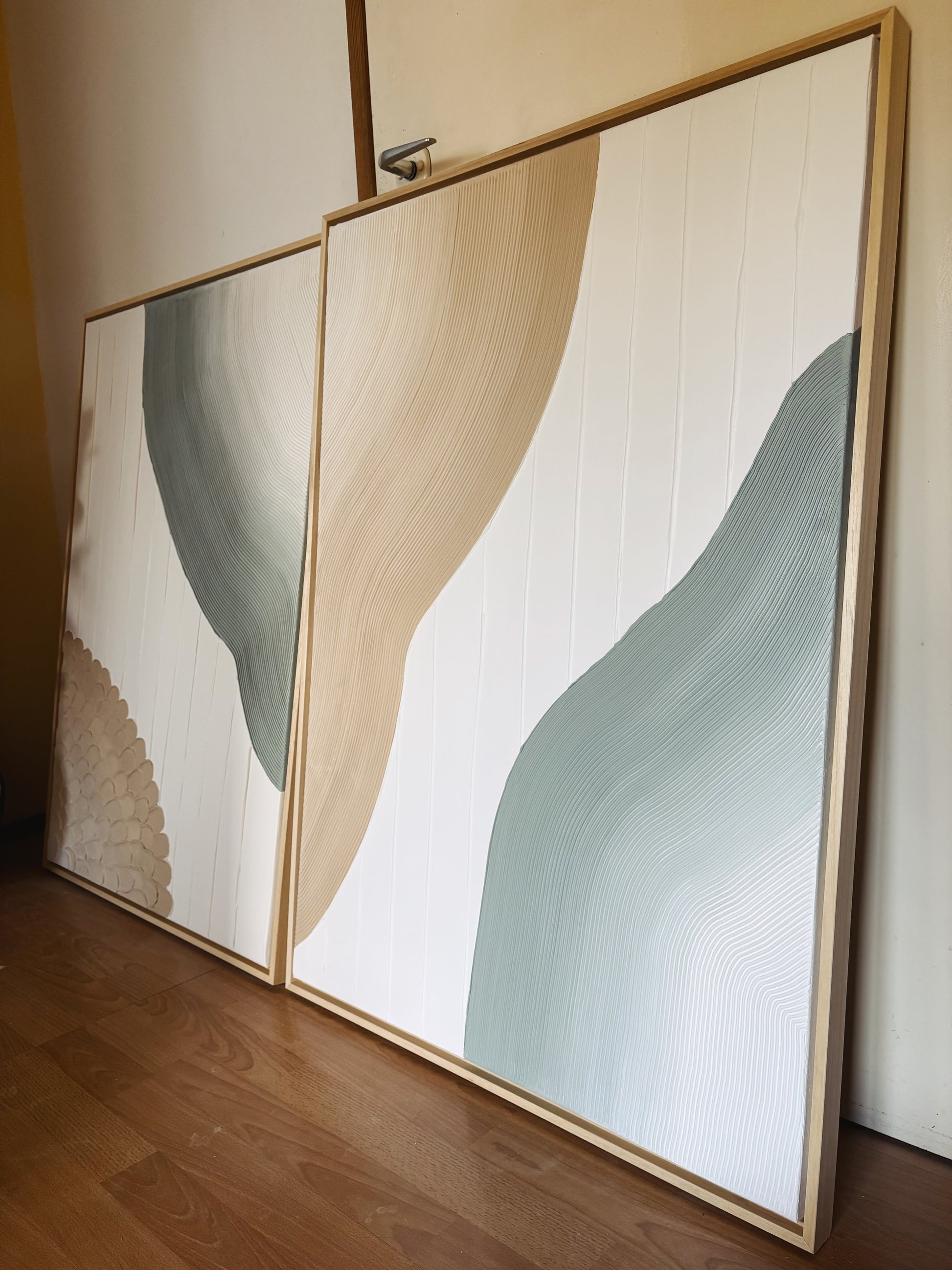 Two abstract wall art pieces with wavy patterns in neutral colors on a wooden floor.