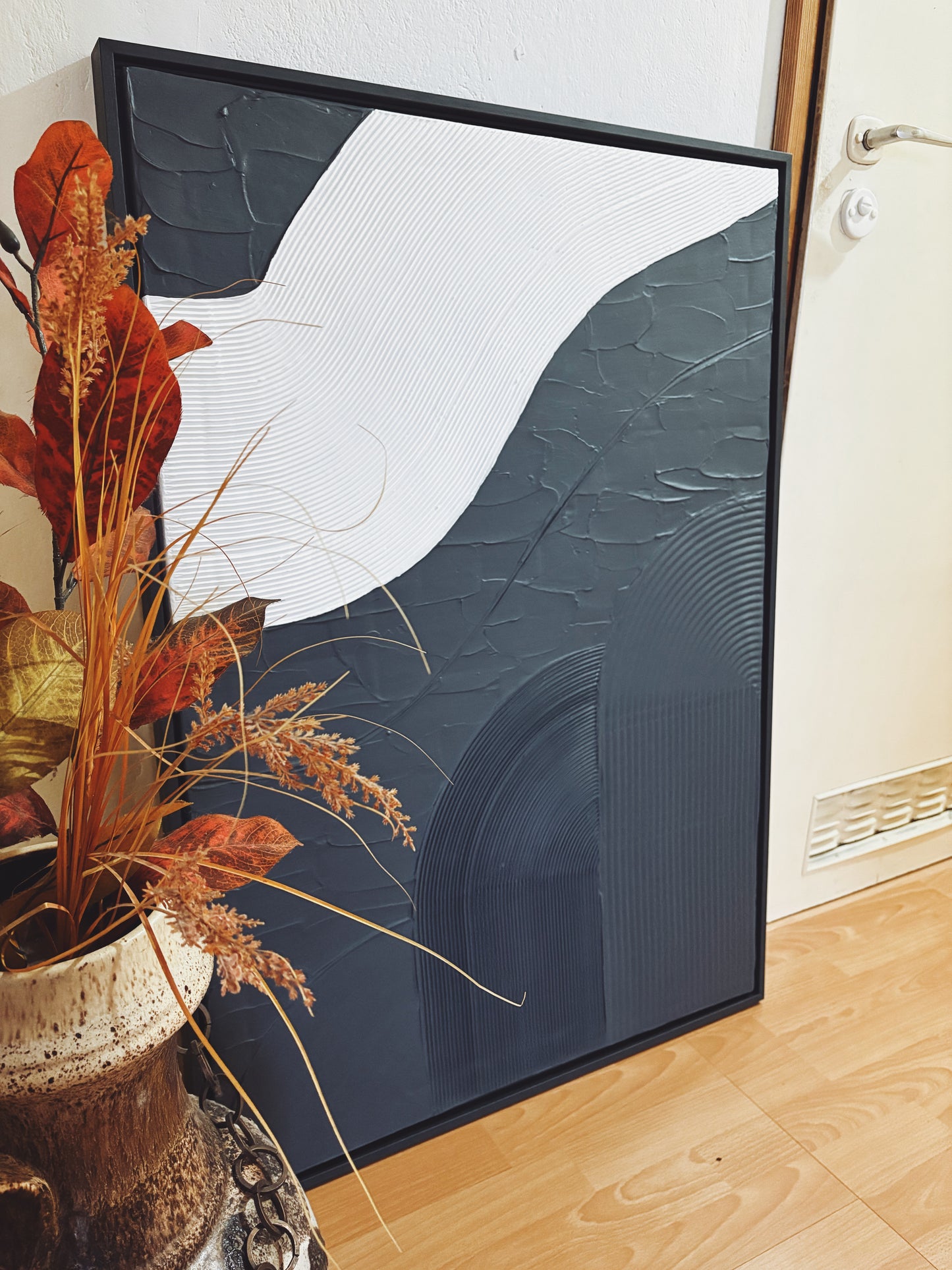 Modern minimalist wall art with abstract design on a white wall, next to a vase with dried plants.