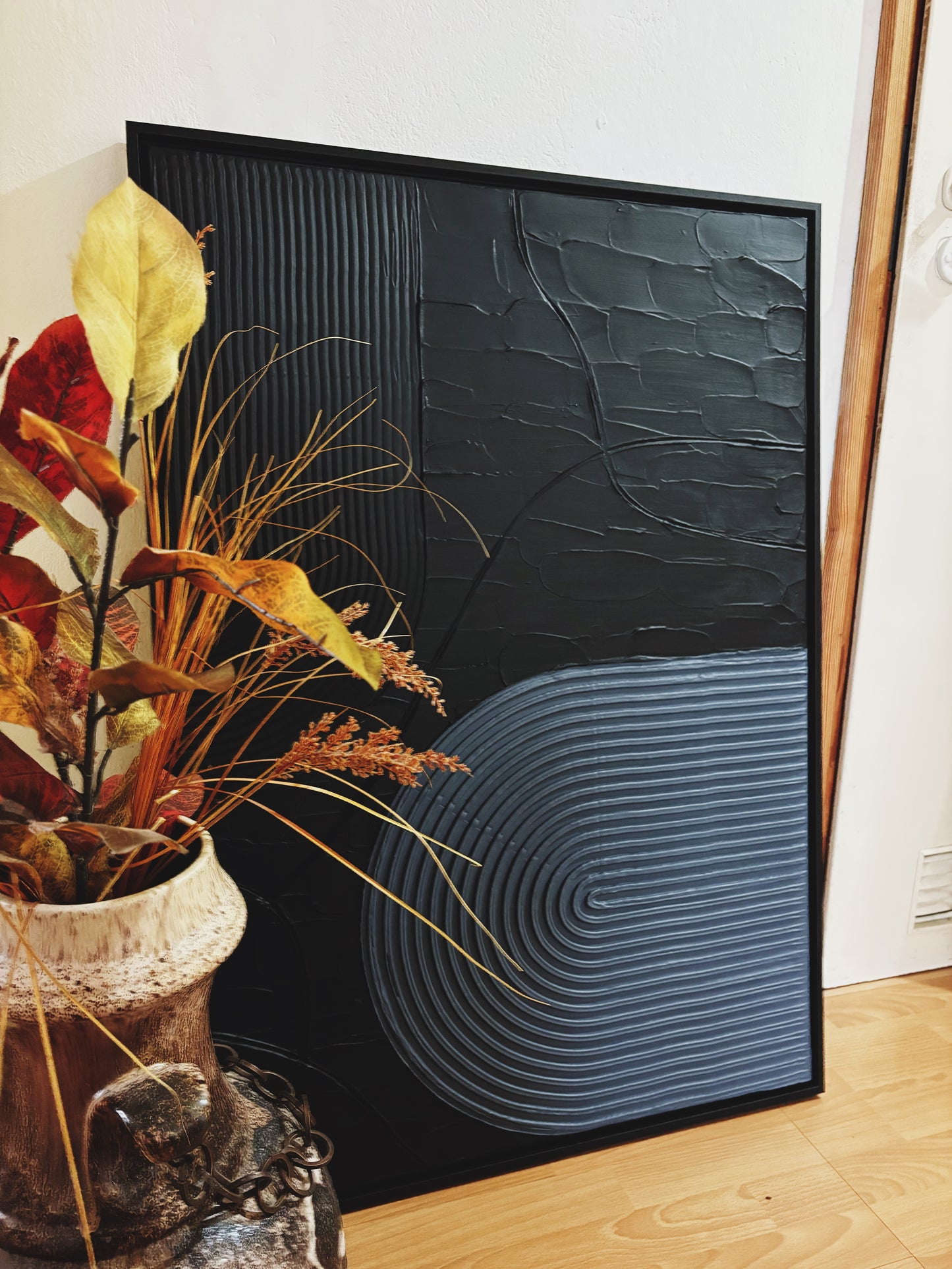 Modern minimalist textured wall art with geometric pattern next to a plant on a wooden floor.