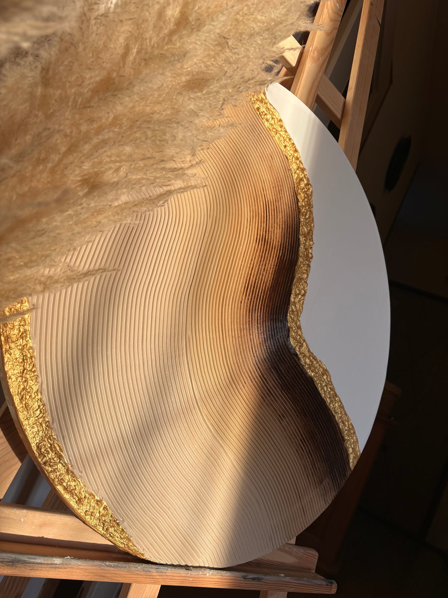 Large wooden disc with gold edge on a wooden stand, surrounded by pampas grass.
