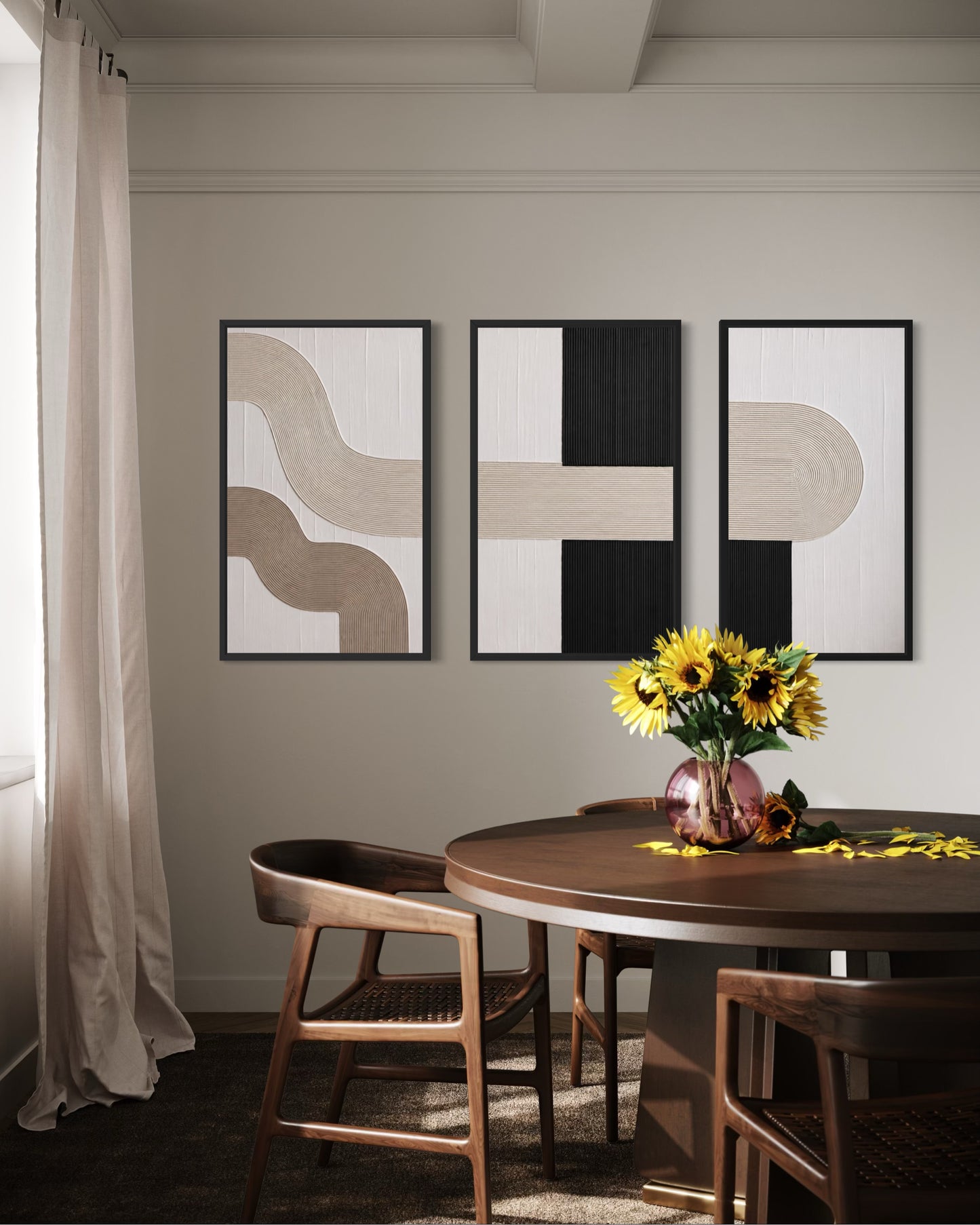 Three framed abstract wall artworks in black, beige, and white hang above a round wooden dining table with chairs and a vase of yellow sunflowers in a softly lit interior.