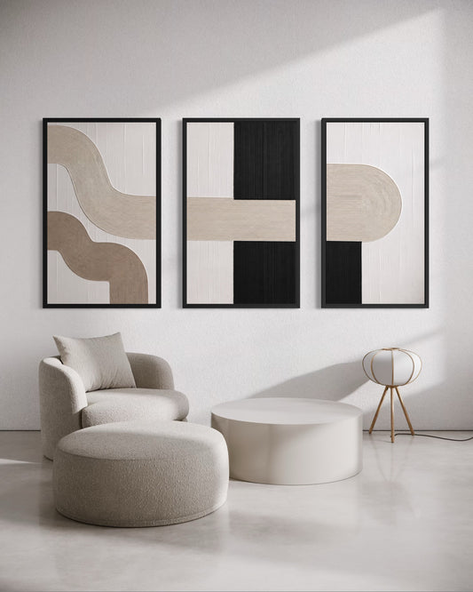 A set of three framed abstract wall art prints with minimalist beige, black, and white geometric shapes, displayed on a white wall above a modern beige armchair, round ottoman, and white coffee table.