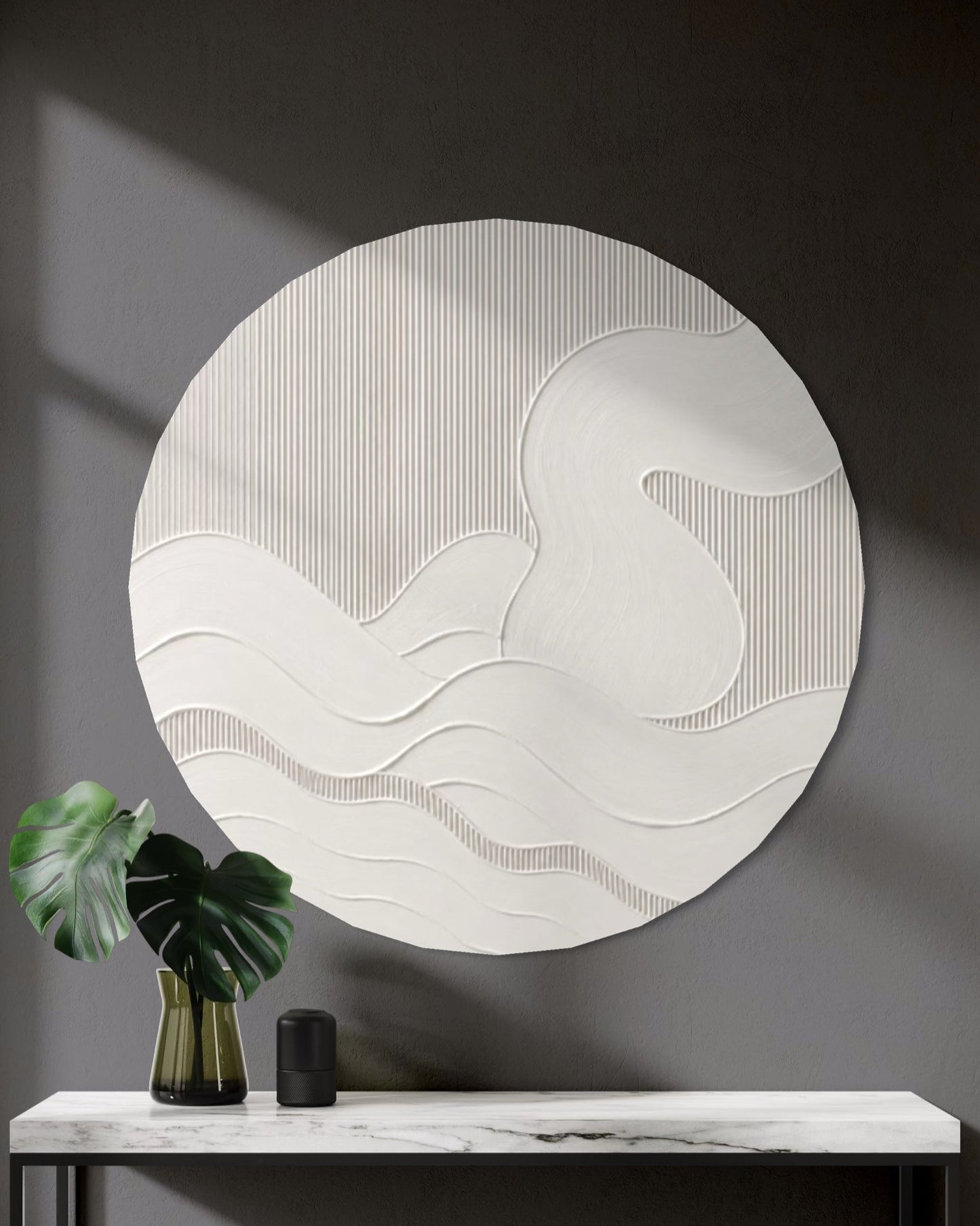 A large circular white textured wall art piece with wavy abstract lines hangs above a marble console table in a modern dark gray entryway. A green monstera leaf in a gold vase and a small black object sit on the table.
