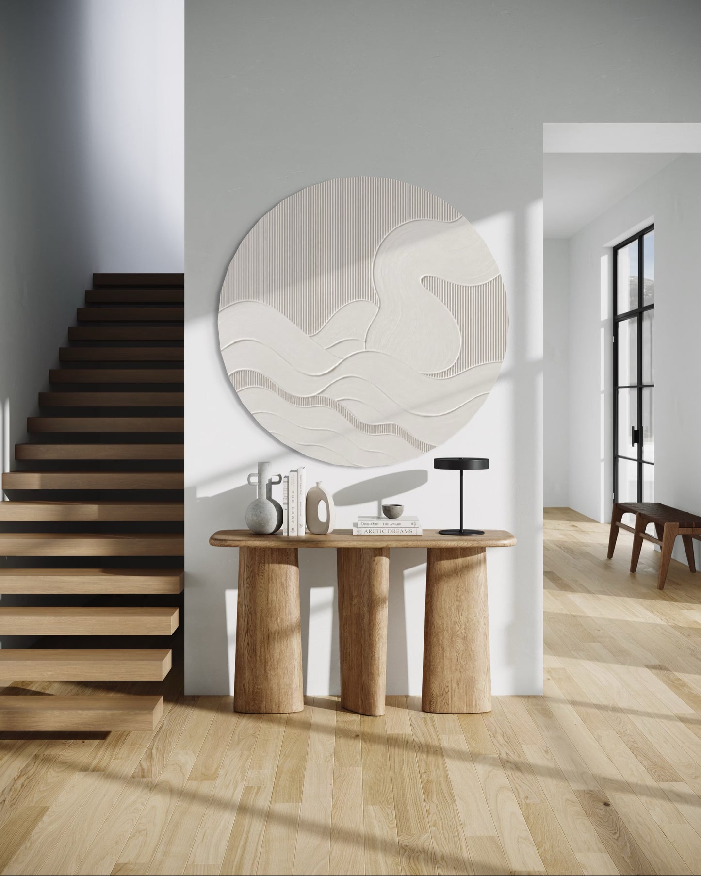 A modern interior scene with a light wood staircase on the left, a wooden console table in the foreground, and a large circular wall art piece featuring abstract flowing white wave patterns on a warm beige background. The room has light gray walls and natural sunlight casting shadows across the floor.