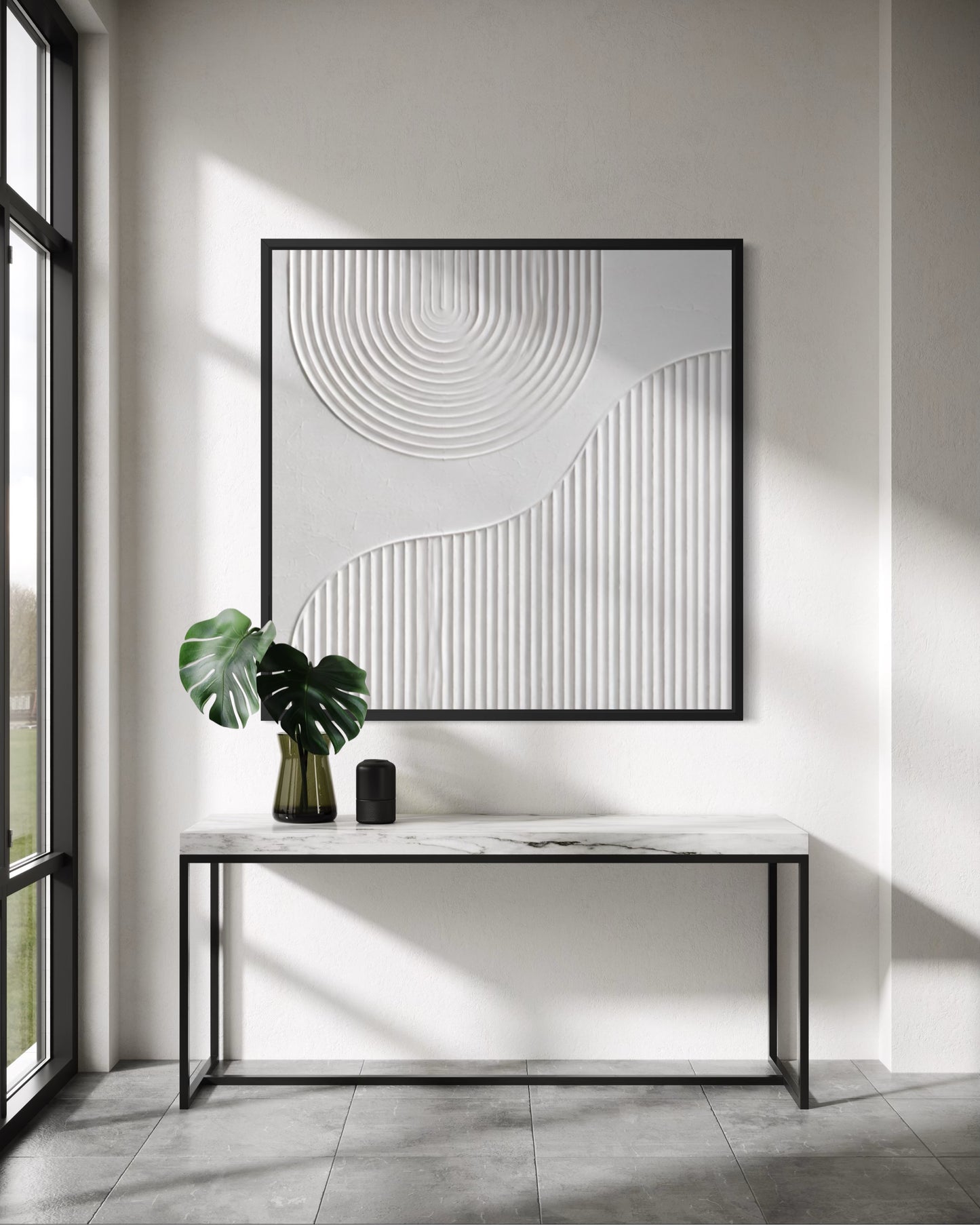 A large square framed wall art piece with abstract white line and circular textured patterns hangs above a marble-top console table in a bright modern room. A green monstera plant and a small black object sit on the table, with sunlight casting strong shadows across the wall and floor.