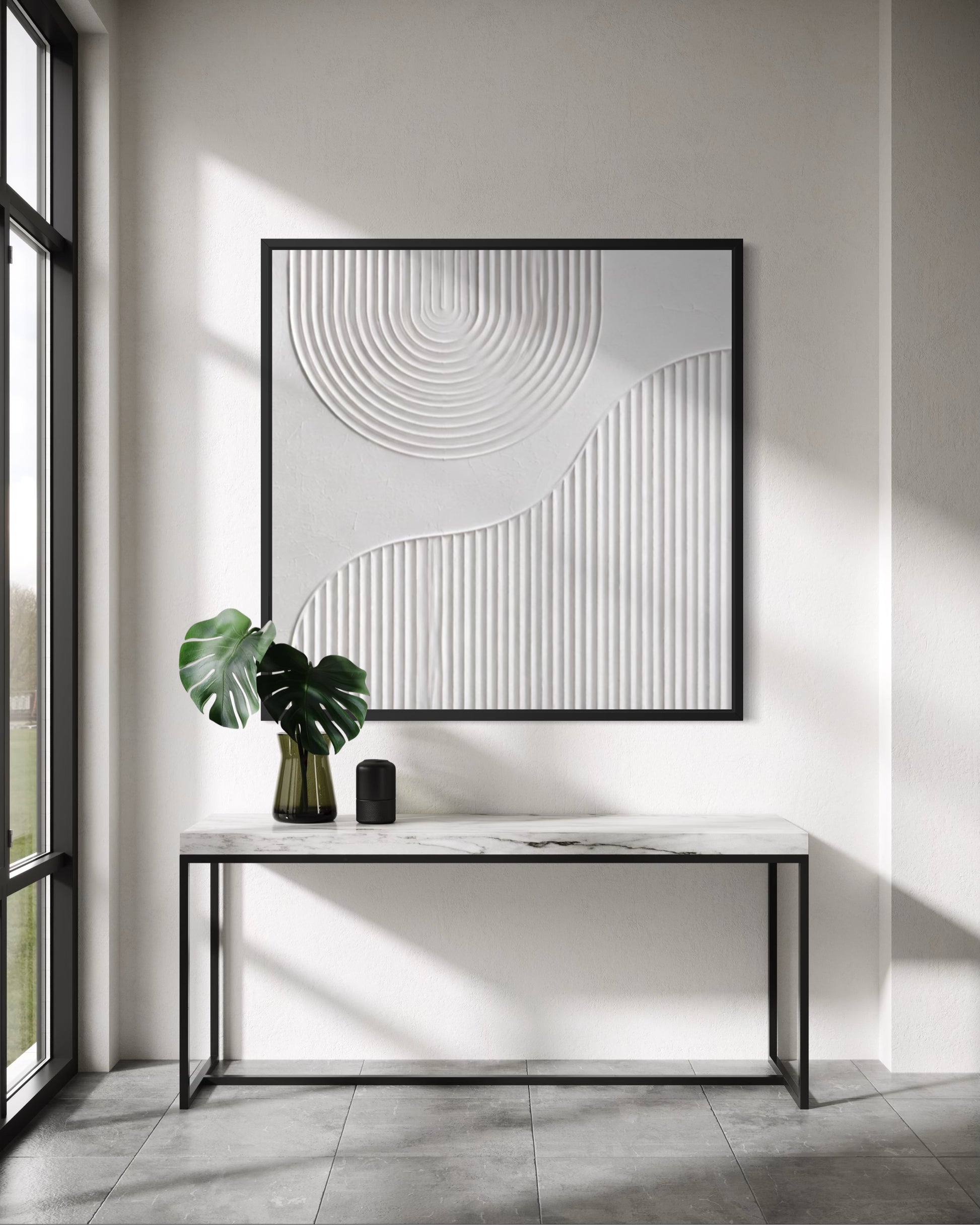 A large square framed wall art piece with abstract white line and circular textured patterns hangs above a marble-top console table in a bright modern room. A green monstera plant and a small black object sit on the table, with sunlight casting strong shadows across the wall and floor.