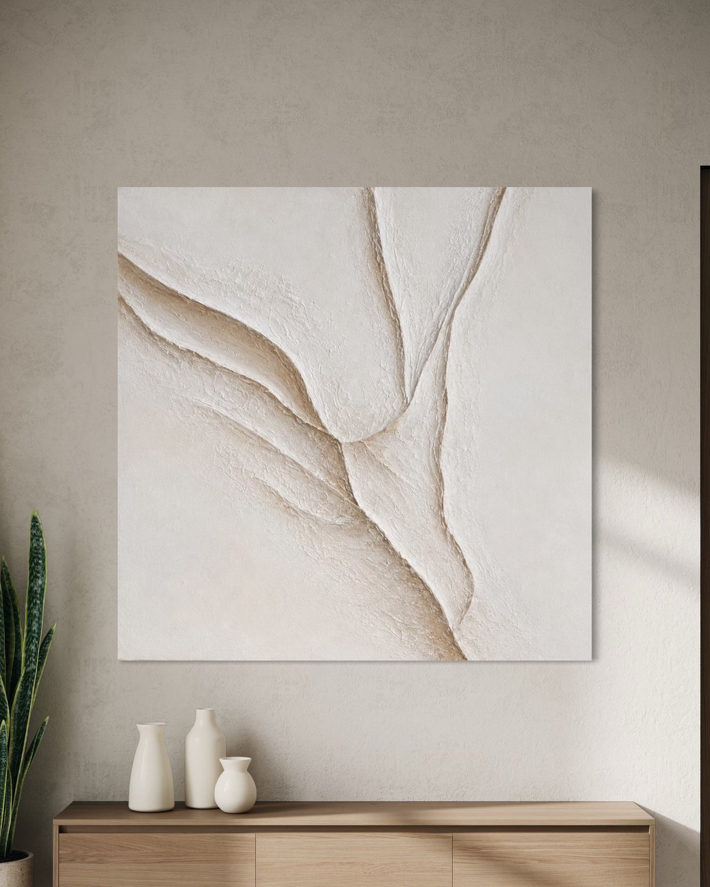 Large square abstract canvas wall art in warm off-white and beige tones, displayed above a light wood console in a modern minimalist interior with a potted snake plant and neutral rug.