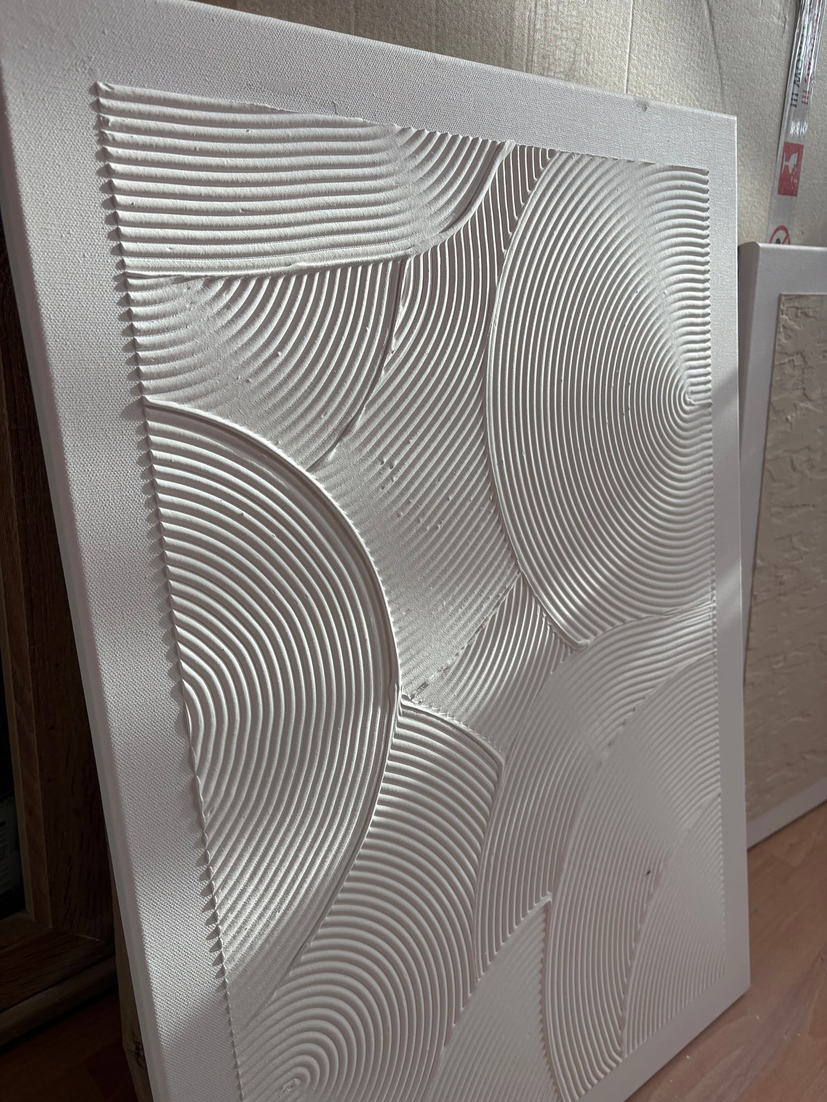 A large square white textured wall art panel with abstract concentric wave and oval patterns, leaning indoors against a light wall.