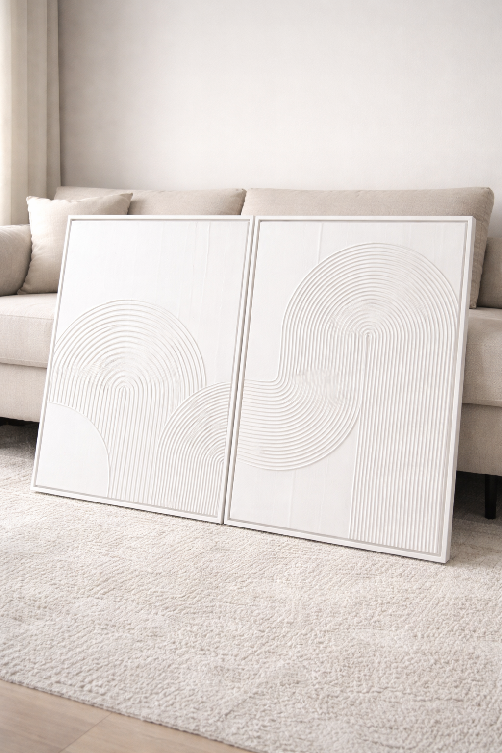 Two white abstract art pieces on a light gray rug with a neutral-colored sofa in the background.