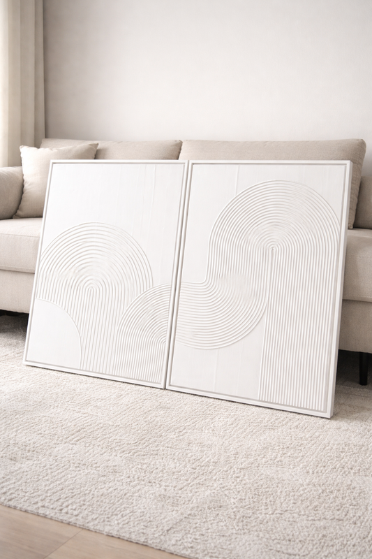 Two white abstract art pieces on a light gray rug with a neutral-colored sofa in the background.